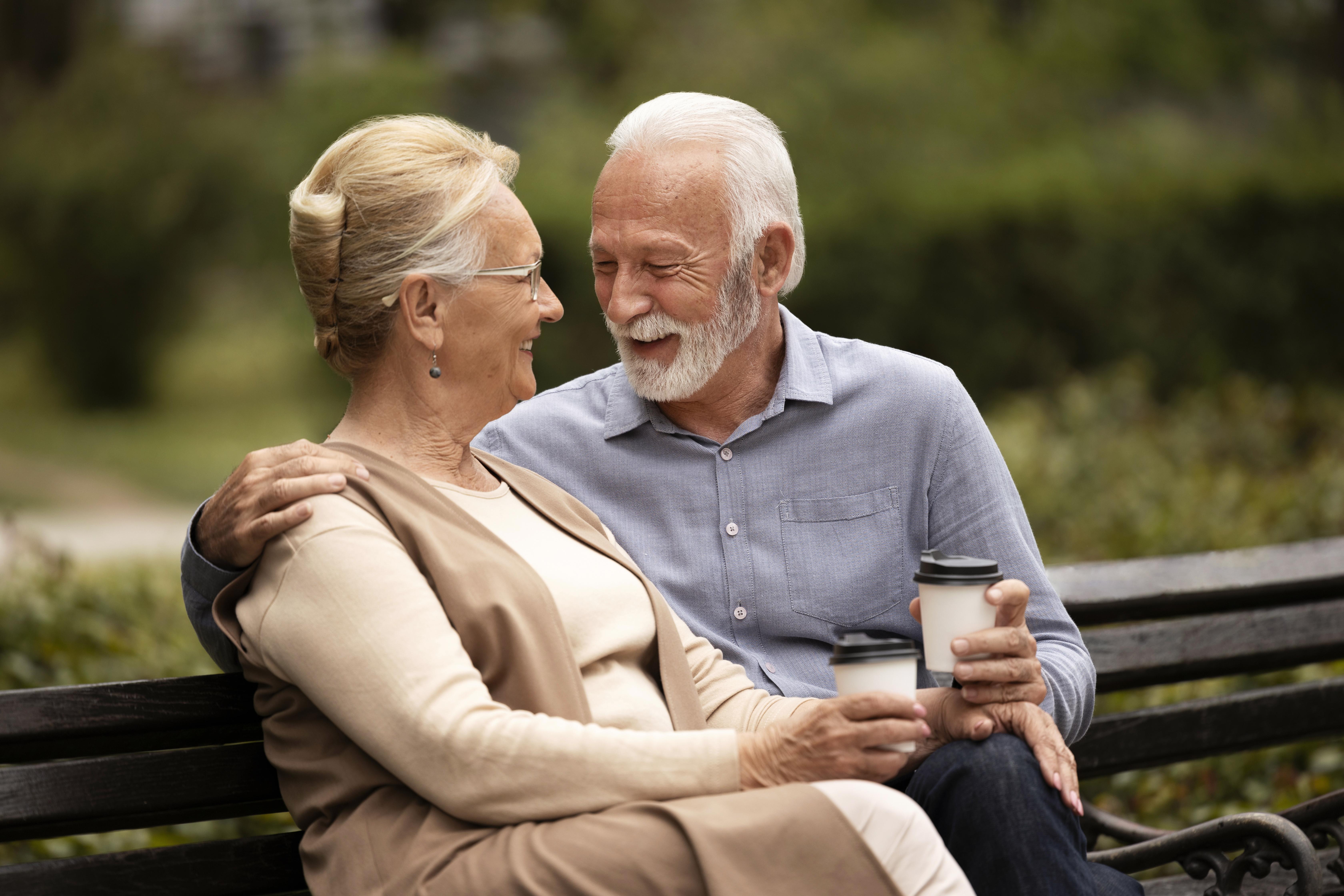 An elderly couple looking at each other, holding coffee cups and sitting on a bench at a park